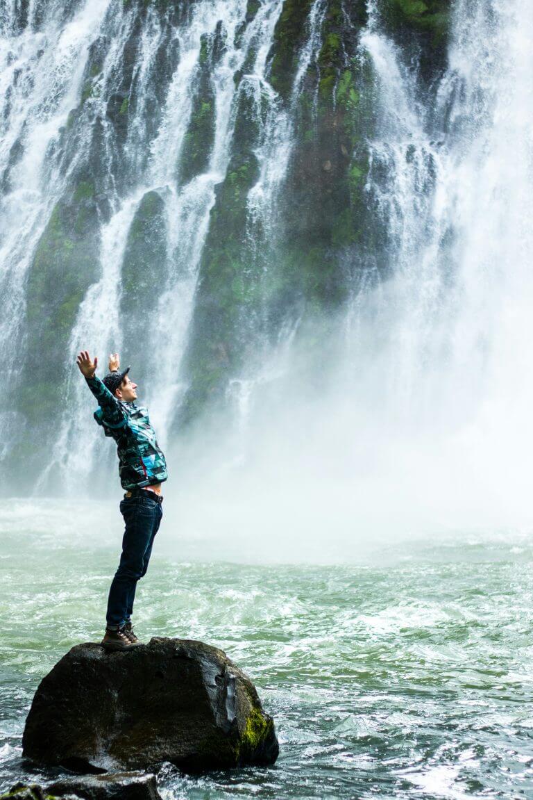 Jeune-homme levant les bras au ciel, sur un rocher dans une rivière, devant une chute d’eau